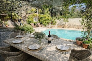 Un espacio exterior para comer con piscina, mobiliario de mimbre y una botella de vino.