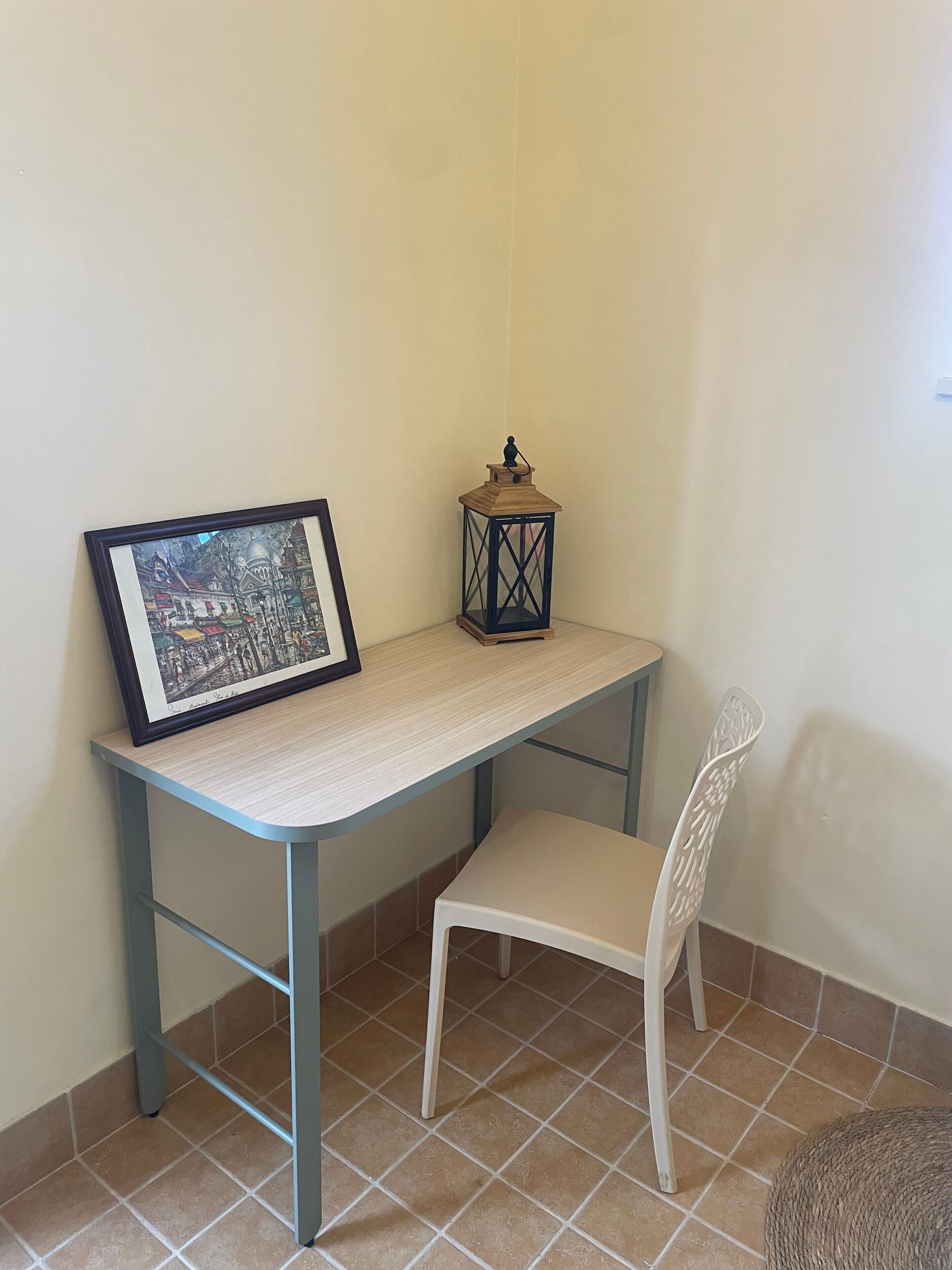 Une pièce d’angle avec un tableau encadré posé sur un bureau de couleur claire, une petite lanterne et une chaise blanche.