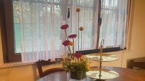 Un tavolo rotondo in legno con un vaso di fiori in vetro, un supporto decorativo in metallo e una finestra con tende leggere.