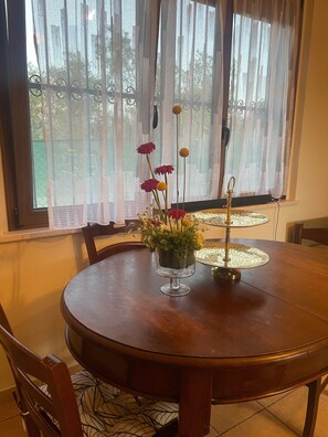 A round wooden dining table with a glass vase of flowers, a decorative metal stand, and a window with sheer curtains.
