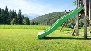 A playground with a green slide and a wooden swing set in a grassy field with mountains in the background. - Apartment “Morgenrot” with Mountain View, Shared Terrace, and Wi-Fi (Ritten)