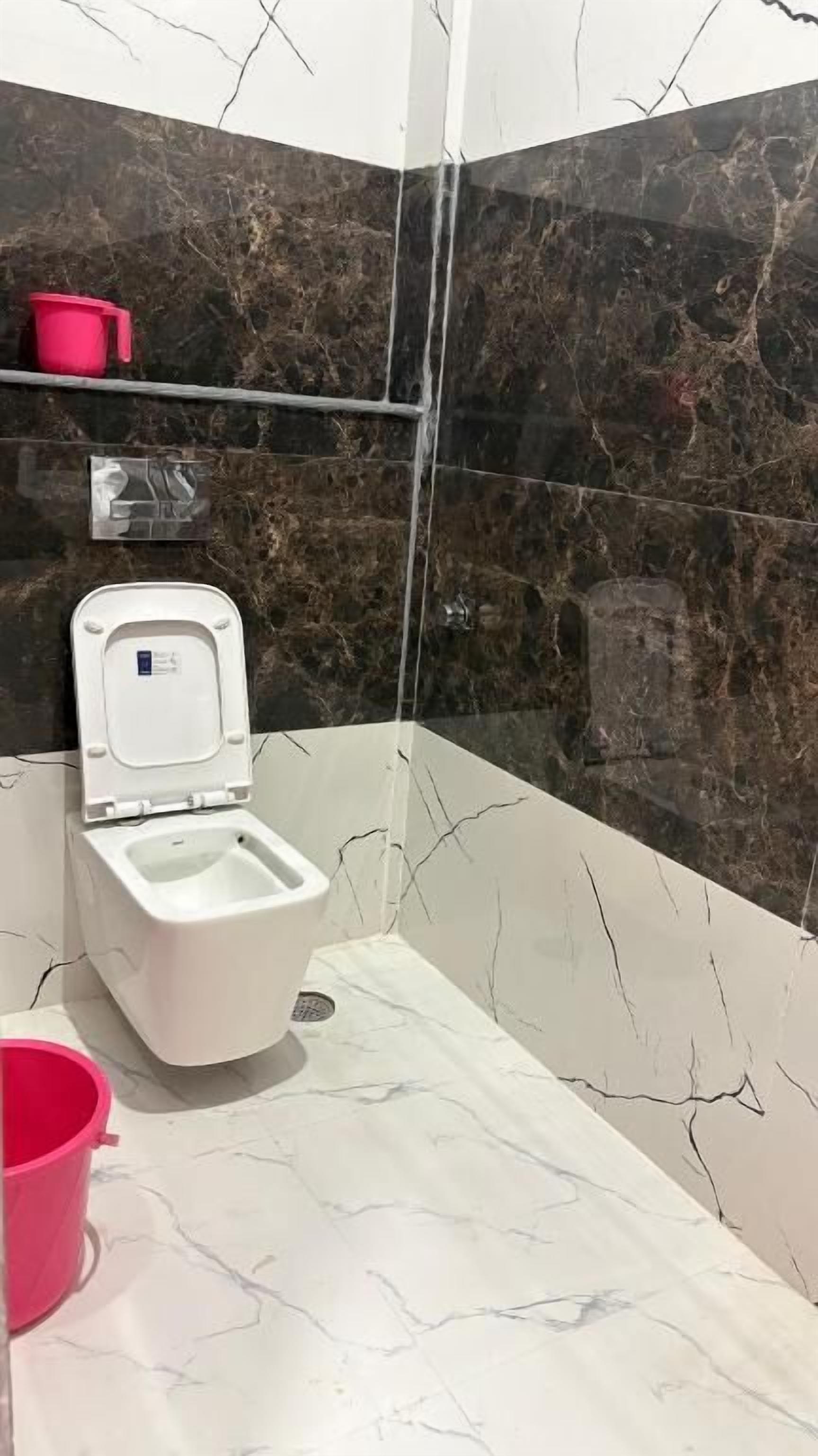 A bathroom with a toilet, a hand dryer, a pink bucket, and a marble-patterned wall.