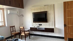 A room with a TV mounted on a wall, a dining table with chairs, and a wooden cabinet. - Shree Laxmi Banquet & Hotel (Darbhanga)