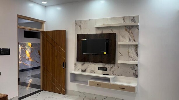 A modern living room with a TV mounted on a marble wall, wooden door, and a small TV stand with drawers. - Shree Laxmi Banquet & Hotel (Darbhanga)