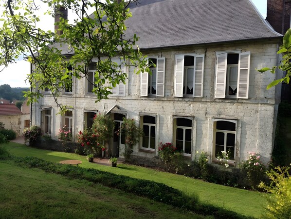 A stone building with multiple windows and shutters, surrounded by a well-maintained garden with various plants and flowers.