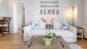 Una sala de estar con sofá, mesa de centro y elementos decorativos, incluyendo una tabla de remo de pared y la palabra 'ALOHA'.