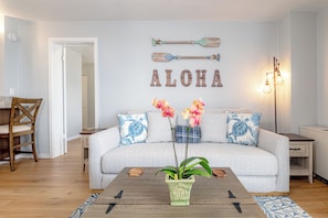 Una sala de estar con sofá, mesa de centro y elementos decorativos, incluyendo una tabla de remo de pared y la palabra 'ALOHA'.