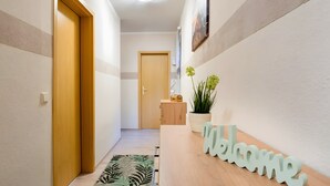 Un couloir avec une porte en bois, une pancarte de bienvenue, une plante et un interrupteur.