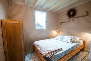 Makuuhuoneessa on puinen sänky, ikkuna, ovi ja seinälle kiinnitetty hylly, jonka päällä on koristeellinen seppele.