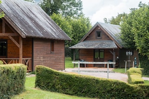 Deux cabanes en bois avec des toits inclinés, une terrasse en bois et un jardin bien entretenu agrémenté de haies.
