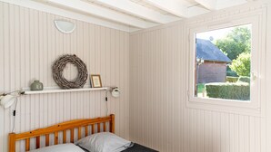 Un dormitorio con cabecera de madera, cama con dos almohadas, estante en la pared con un adorno y un objeto enmarcado, lámpara de noche y ventana con vista a una casa y follaje.