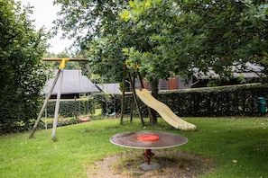 Een speelplaats met een glijbaan en schommels, omringd door een haag en bomen.