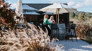 Een terras met tafels en stoelen, een grote parasol, en een rustiek gebouw op de achtergrond.