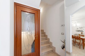 Una porta in legno con un pannello di vetro satinato, una scala che conduce a una sala da pranzo con tavolo e sedie, e una parete con un cartello decorativo.