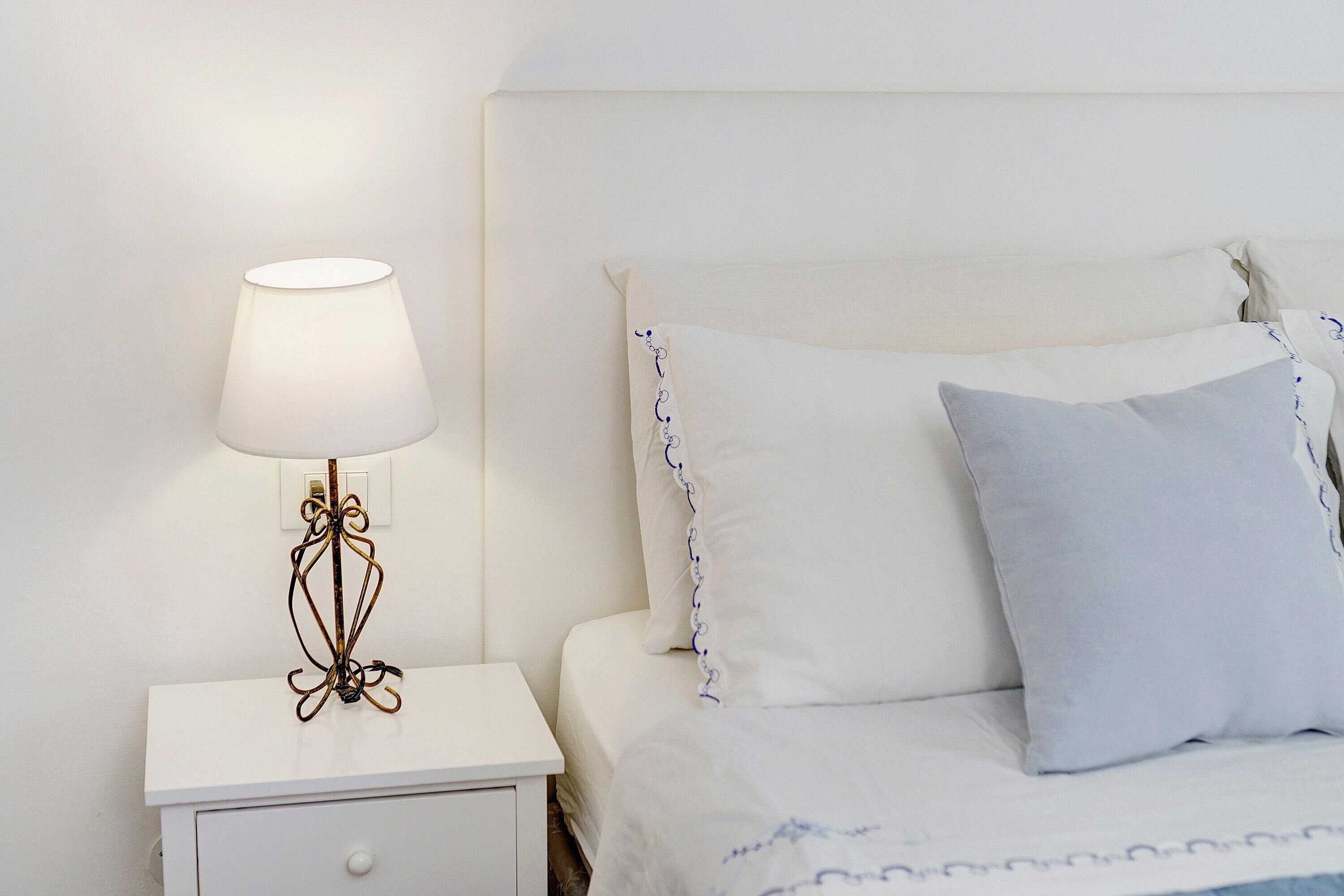 Un lit avec une literie blanche et un seul oreiller bleu, une table de chevet avec une lampe et une commode blanche avec un tiroir.