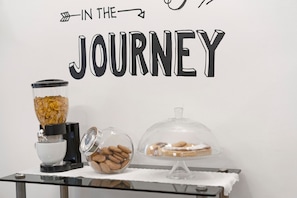 A breakfast setup with a cereal dispenser, a bowl of cookies, and a covered dessert display on a table against a wall with the text 'IN THE JOURNEY'.