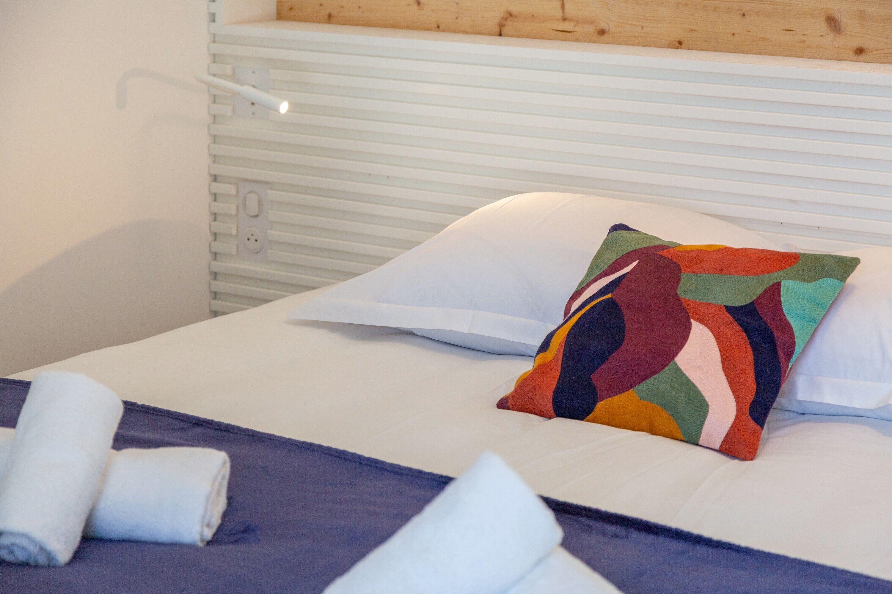 A neatly made bed with a colorful pillow, a rolled white towel, and a white headboard.