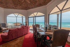 A spacious living room with a red sofa, dining table with chairs, and large arched windows overlooking the ocean. - Cerquita del Mar – Beachfront, Rocky Point (Puerto Peñasco)