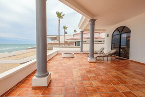 Ein überdachter Außenbereich mit gefliestem Boden, einer Feuerstelle, Liegestühlen und Blick auf Strand und Meer.
