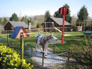 Un área cercada con una escultura de vaca, un parque infantil con columpios y toboganes, y un edificio con techo inclinado.