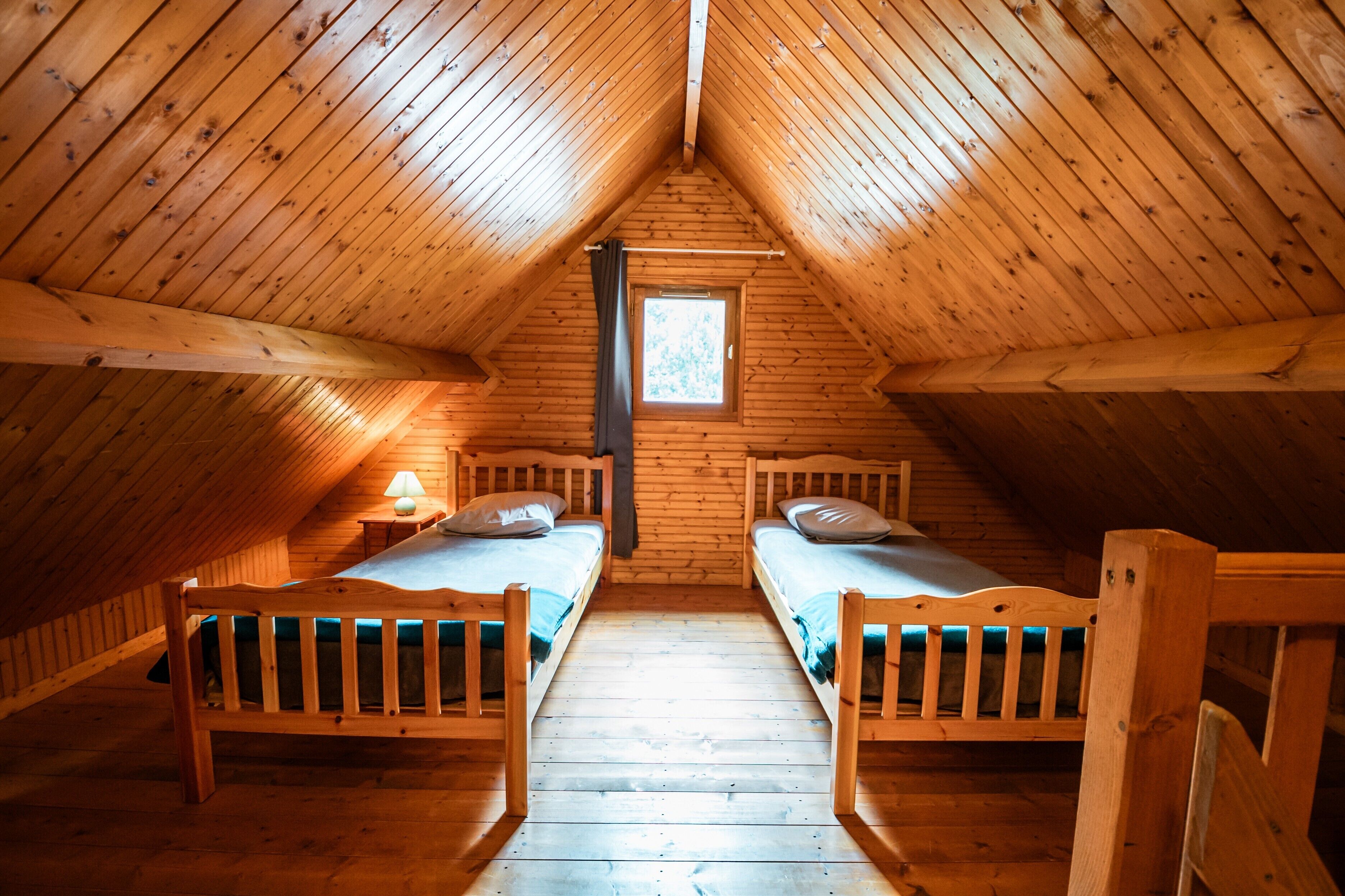 A room with wooden walls and a sloped ceiling, two beds, a window, and a nightstand with a lamp.