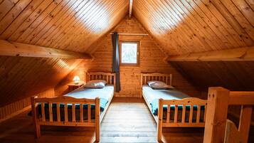 A room with wooden walls and a sloped ceiling, two beds, a window, and a nightstand with a lamp.