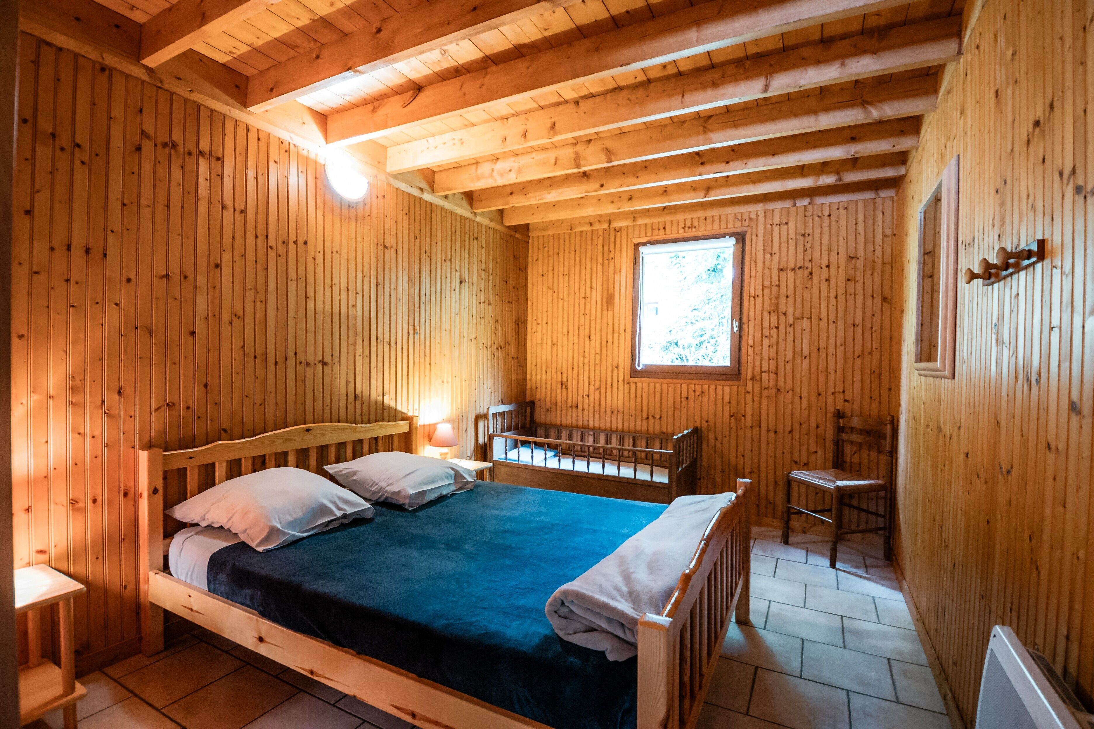 Un dormitorio de cabaña de madera con una cama, una cuna, una silla y una ventana.