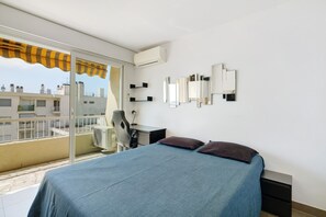 A bedroom with a bed, a desk, a chair, and a balcony with a view of buildings. - Apartment 'T2 Stunning Sea View' with Sea View, Wi-Fi and Air Conditioning (Cagnes-sur-Mer)
