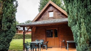 A wooden cabin with a covered patio, a dining table with chairs, and a small table with a bench. - Chalet Noël des Champs for 8 with Shared Pool, Terrace, and Wi-Fi (Livarot-Pays-d'Auge)