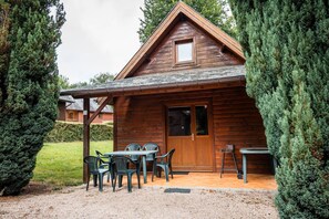 A wooden cabin with a covered patio, a dining table with chairs, and a small table with a bench. - Chalet Noël des Champs for 8 with Shared Pool, Terrace, and Wi-Fi (Livarot-Pays-d'Auge)
