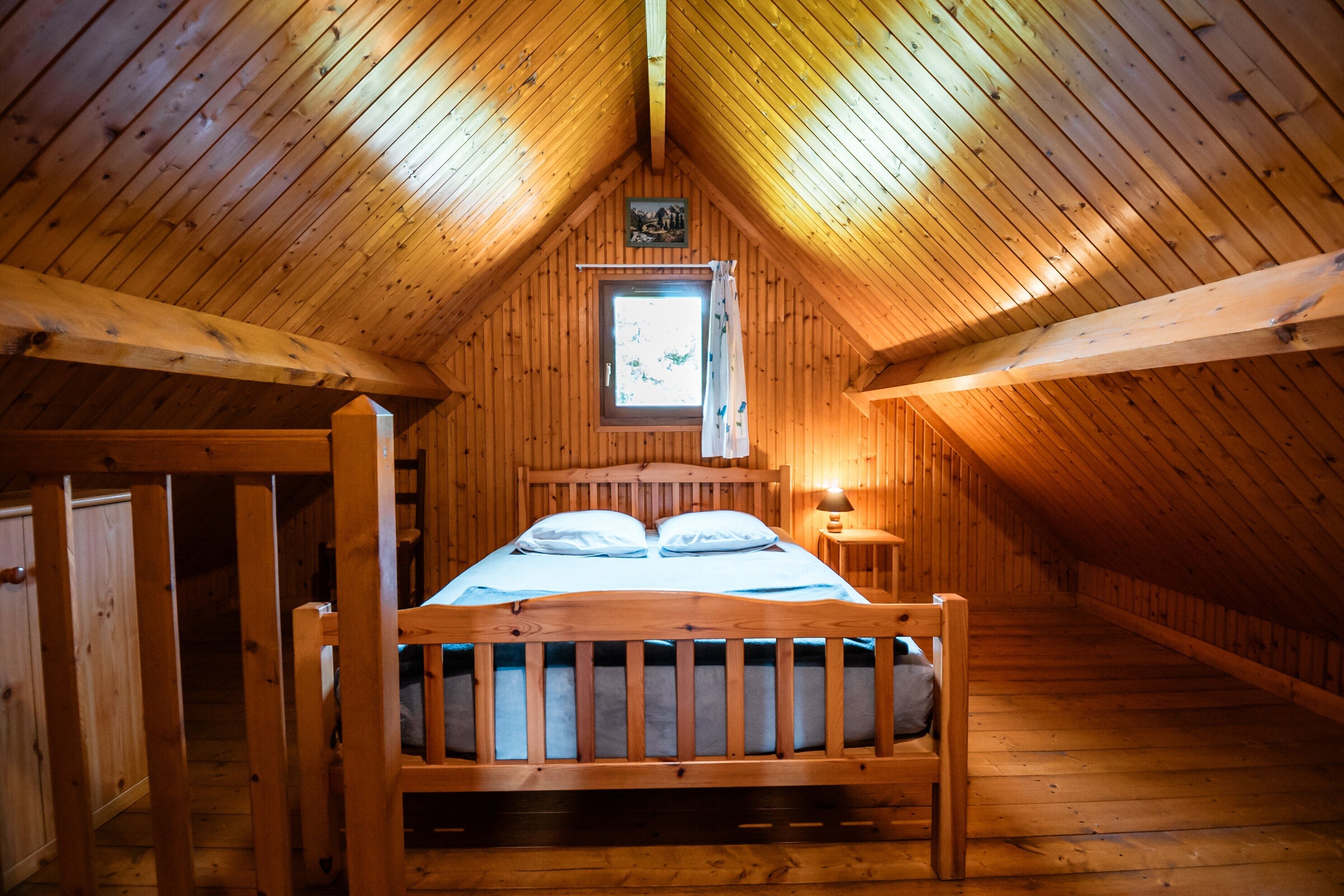 Quarto de cabana de madeira com cama, janela com cortinas e um pequeno quadro pendurado na parede.