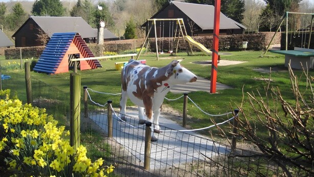 A fenced area with a sculpture of a cow, a playground with swings and slides, and a building with a sloped roof. - Chalet Noël des Champs for 8 with Shared Pool, Terrace, and Wi-Fi (Livarot-Pays-d'Auge)