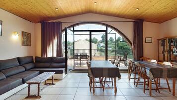 Een ruime woonkamer met een houten plafond, een grote bank, een eettafel met stoelen en uitzicht buiten via een glazen deur.