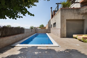 A rectangular swimming pool with a tiled deck, a closed garage door, and a stack of lounge chairs.