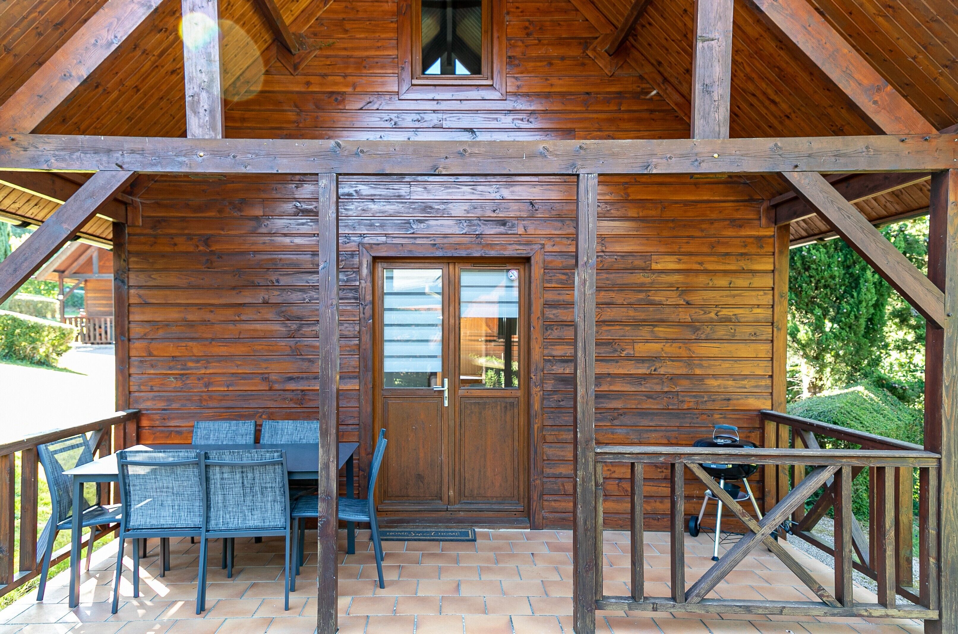 A wooden cabin with a patio area featuring a table and chairs, a grill, and a door with glass panels.