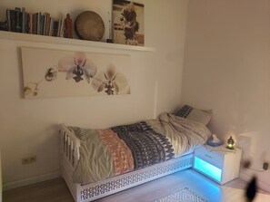 A bedroom with a bed, a nightstand, a wall-mounted shelf with books and decorative items, and a wall art piece. - Zen Moroccan Cozy Guest Room (Antwerpen)