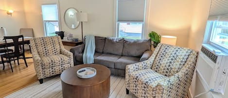 A living room with a sofa, two patterned armchairs, a coffee table, a dining table with chairs, a mirror, and a window with blinds.