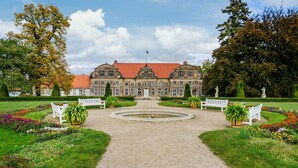 Een groot, historisch gebouw met een symmetrische gevel, een fontein in het midden en een verzorgde tuin met bankjes.
