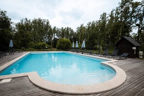 A rectangular swimming pool with a wooden deck, surrounded by lounge chairs and umbrellas, and a small cabin-like structure in the background. - Domaine de Guillaume, Chalet Frequin 4p with Shared Pool, Wi-Fi (Livarot-Pays-d'Auge)