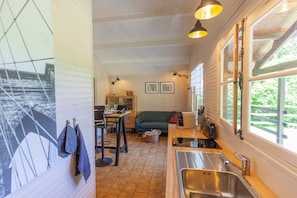 A compact kitchen with a sink, stove, and dining area, a sofa, and a window with a view of greenery. - Domaine de Guillaume, Chalet Frequin 4p with Shared Pool, Wi-Fi (Livarot-Pays-d'Auge)