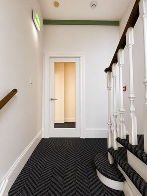 Un escalier avec des balustrades blanches et une moquette à motifs sombres menant à une porte dont l’intérieur est éclairé.