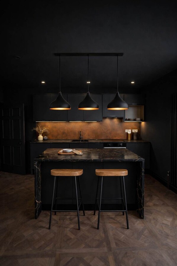 A modern kitchen with a dark color scheme, three pendant lights, a central island with stools, and a herringbone wood floor. - The Bridge York - Stunning River Front Location (York)