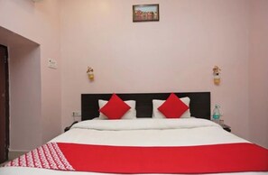 Habitación de hotel con una cama, dos almohadas rojas, una colcha blanca y roja, un cuadro en la pared y una botella de agua en la mesita de noche.