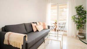 Ett modernt vardagsrum med en grå soffa, ett soffbord i glas, en krukväxt och utsikt över utomhusmiljön genom ett fönster med gardiner.