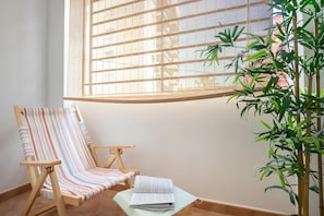 Ein Zimmer mit großem Fenster, einem gestreiften Sessel, einem kleinen Tisch mit einem Buch und einer Bambuspflanze.