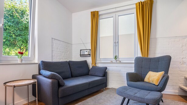 Ein modernes Wohnzimmer mit einem grauen Sofa, einem passenden Sessel, einem kleinen Beistelltisch und einem Fenster mit Vorhängen.