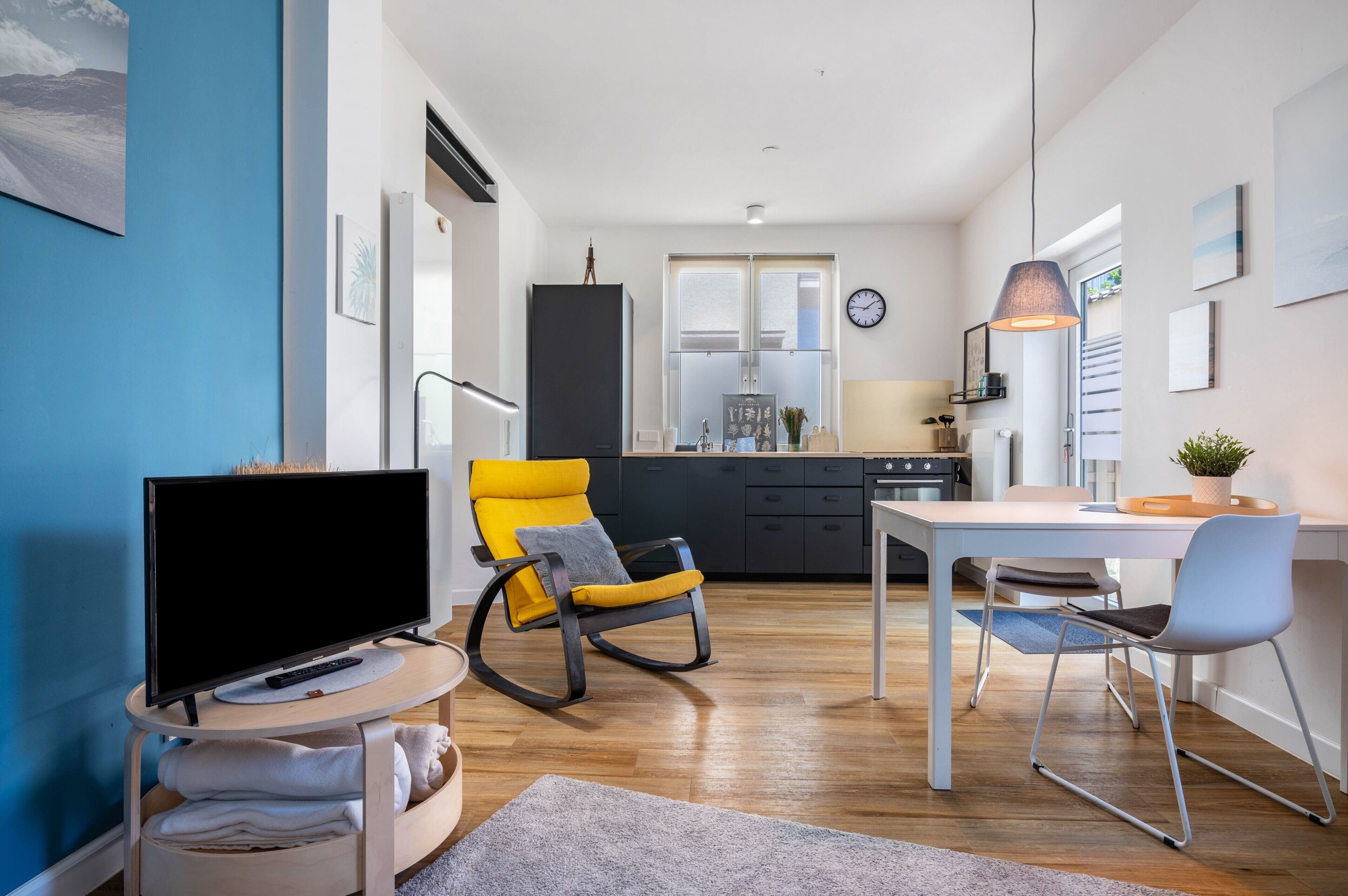 A modern living room with a TV, a yellow rocking chair, a dining table with chairs, a kitchen in the background, and a clock on the wall.
