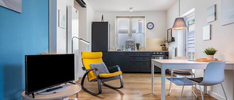 A modern living room with a TV, a yellow rocking chair, a dining table with chairs, a kitchen in the background, and a clock on the wall.