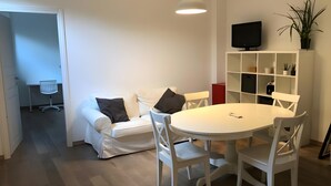 Ein modernes Wohnzimmer mit einem weißen Sofa, einem runden Esstisch, weißen Stühlen, einem Fernseher auf einem Regal und einer Topfpflanze.