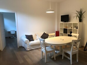 Ein modernes Wohnzimmer mit einem weißen Sofa, einem runden Esstisch, weißen Stühlen, einem Fernseher auf einem Regal und einer Topfpflanze.